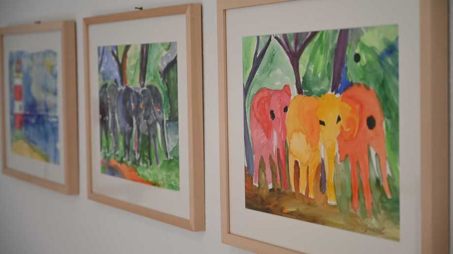 Holz-Bilderrahmen hängen an einer weißen Wand. In den Rahmen sind gemalte, bunte Bilder von Elefanten.