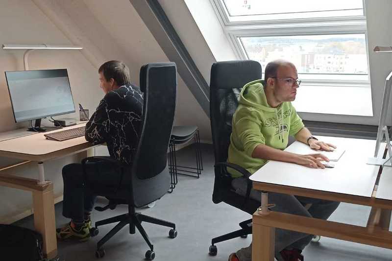 Zwei Menschen sitzen an Schreibtischen vor einem Computerbildschirm