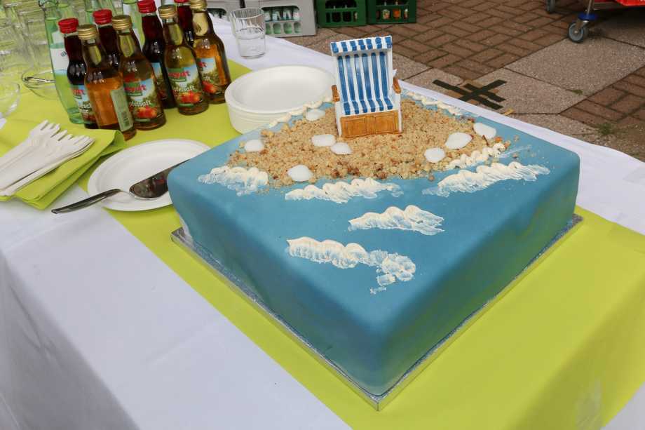 Eine quadratische, blaue Torte. Darauf angedeutete Wellen, die an einen Strand aus Kekskrümeln stoßen. Darauf ein Strandkorb.