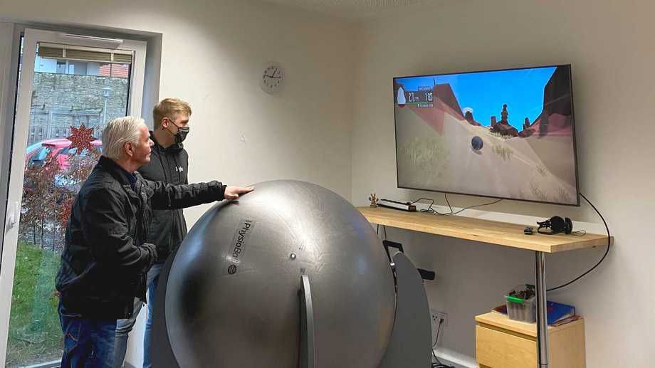Ein Mann steht hinter einem Gymnastikball, der in einem Gestell befestigt ist. An einer Wand hängt ein Fernseher.