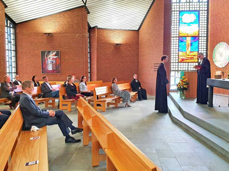 Ein Kirchengebäude mit roten Backsteinwänden und Holz-Sitzbänken. Im Altarbereich zwei Pastoren im Talar.