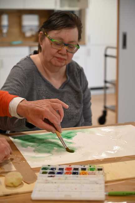 Eine Frau mit Brille sitzt an einem Tisch. Darauf ein Blatt Papier mit grüner Farbe. Die Frau hält einen Pinsel in der Hand. Die Hand einer nicht weiter sichtbaren Person führt ihre Hand.