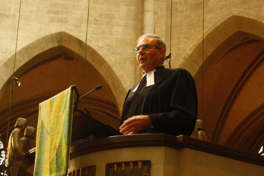 Pastor Ulrich Stoebe trägt einen Talar und steht auf einer Kanzel in einer Kirche.