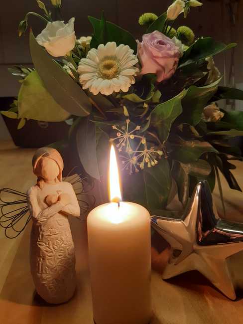Eine brennende Kerze vor einem Blumenstrauß. Daneben ein Engel und ein Stern.
